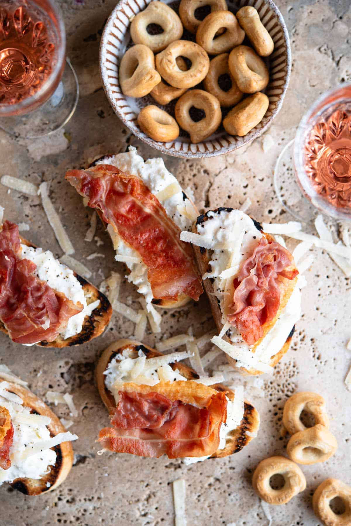 An overhead shot of burrata and Parmigiano crostini on a travertine background topped with prosciutto.