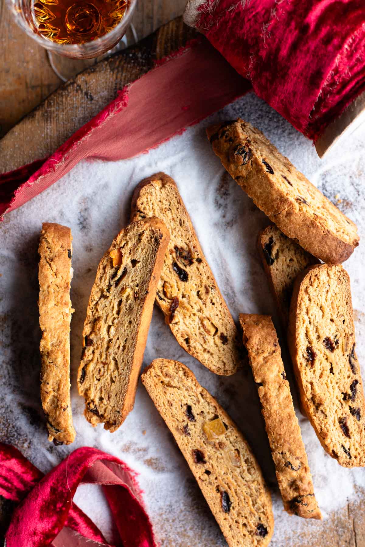 An overhead shot of panettone biscotti on a wooden surface with sugar and a red Christmas ribbon.