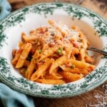 A cropped square image of a bowl of creamy tomato penne pasta on a wooden table.