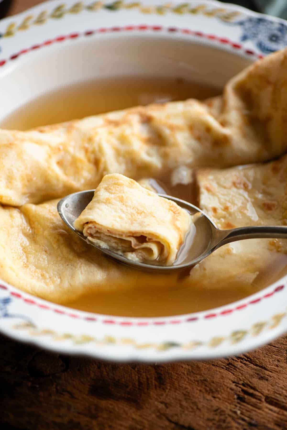 A close up of a spoon in a bowl of broth picking up a spoonful of crepe.