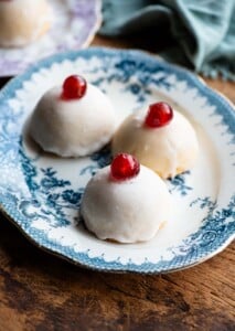 Three Sicilian pastries sitting on a blue patterned plate, the pastries are dome shaped with white icing and a cherry on top.