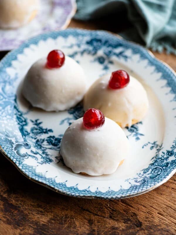 Three Sicilian pastries sitting on a blue patterned plate, the pastries are dome shaped with white icing and a cherry on top.