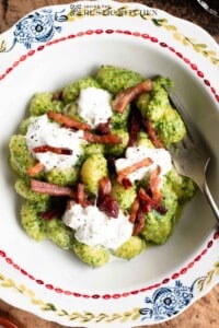 An overhead shot of a bowl of arugula pesto gnocchi topped with burrata cheese and crispy pancetta.
