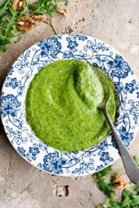 An overhead shot of arugula pesto in a blue and white patterned bowl with a spoon at the side.