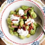 An cropped image of arugula pesto gnocchi in a bowl topped with pancetta and burrata.