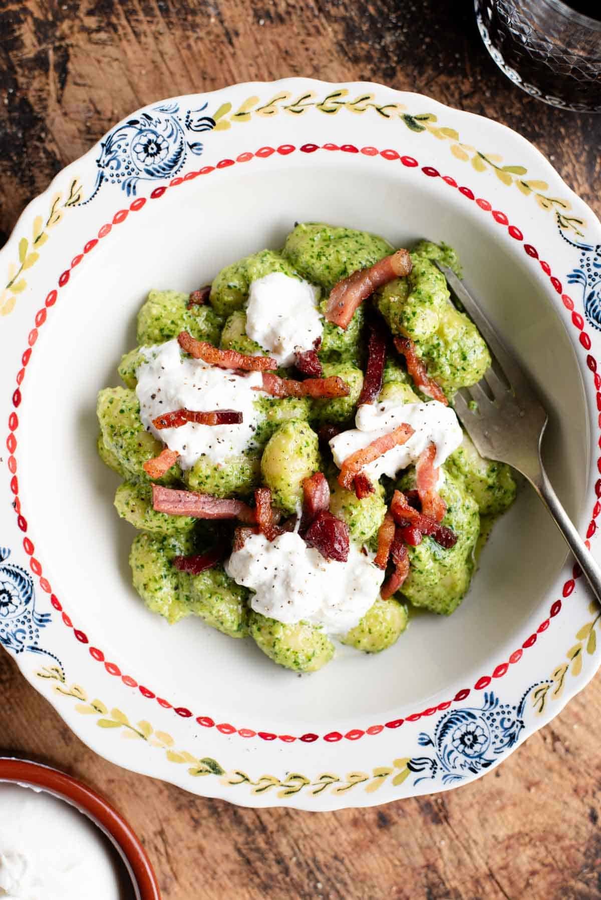 An overhead shot of arugula pesto gnocchi in a bowl topped with crispy pancetta and burrata.