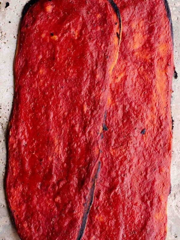 Two long thin and crispy pizzas covered in tomato sauce sitting on a travertine surface.