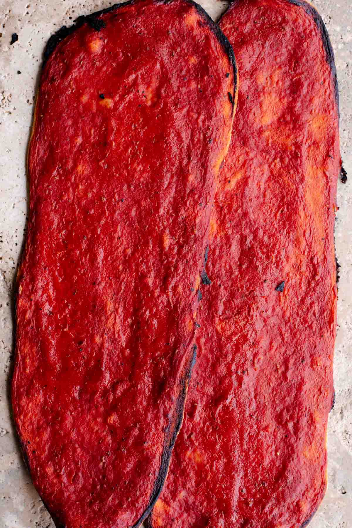 Two long thin and crispy pizzas covered in tomato sauce sitting on a travertine surface.