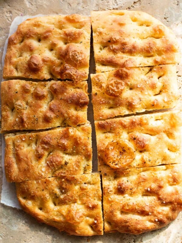 Golden, rustic focaccia bread sliced into rectangular pieces, with a crisp, dimpled crust, a sprinkle of sea salt, and a light seasoning of herbs, resting on a stone surface.