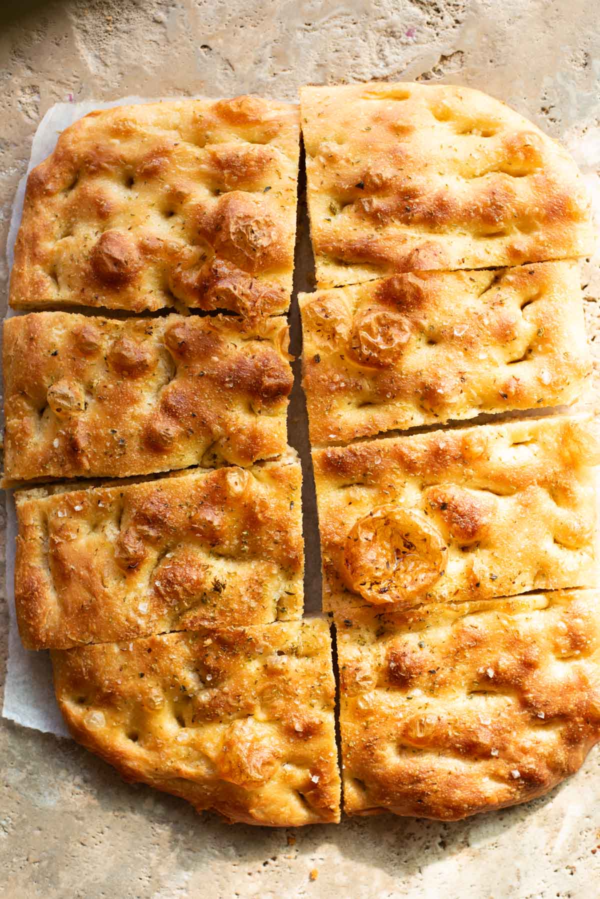 Golden, rustic focaccia bread sliced into rectangular pieces, with a crisp, dimpled crust, a sprinkle of sea salt, and a light seasoning of herbs, resting on a stone surface.