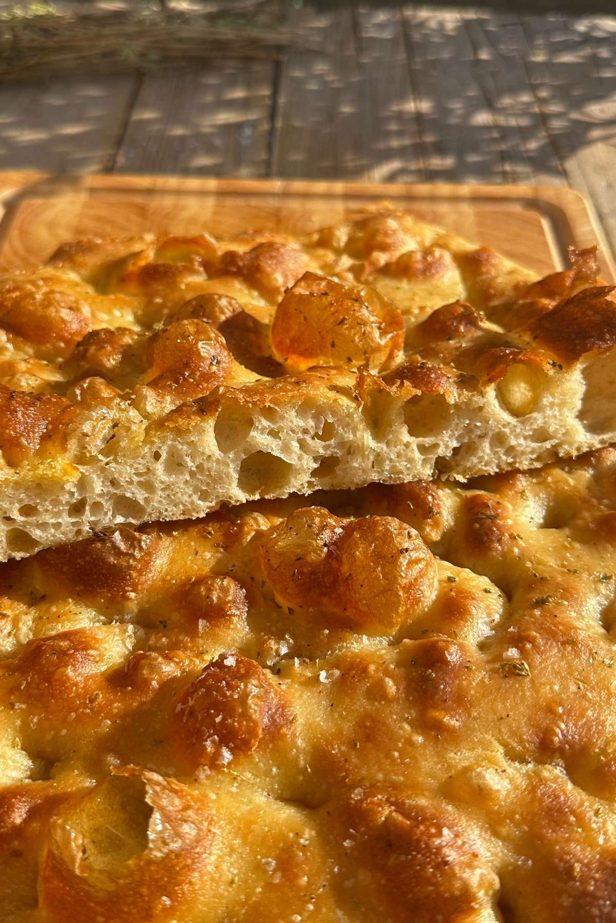 A golden and airy focaccia cut in half to show it's bubbly texture sitting on a wooden chopping board.