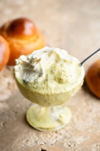A close up of pistachio granita in a glass serving bowl topped with whipped cream and a brioche bun behind it.