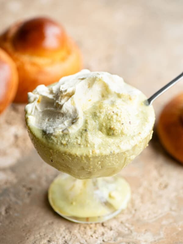 A close up of pistachio granita in a glass serving bowl topped with whipped cream and a brioche bun behind it.