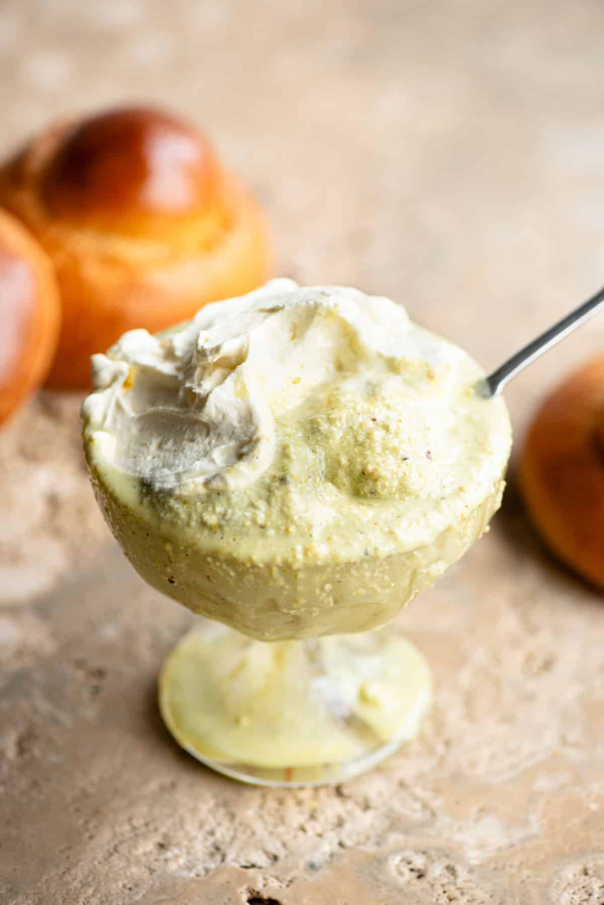 A close up of pistachio granita in a glass serving bowl topped with whipped cream and a brioche bun behind it.