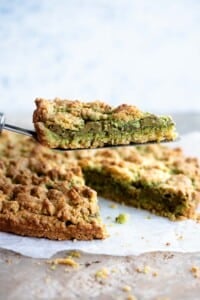 A cake slice holding up a slice of pistachio crumb cake showing a layer of pistachio in the middle.