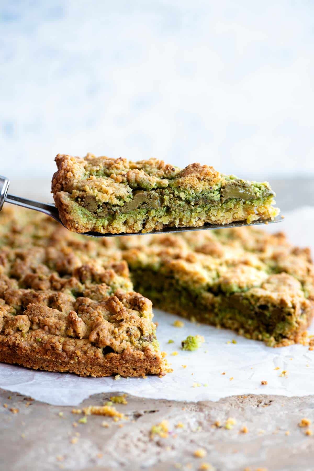 A cake slice holding up a slice of pistachio crumb cake showing a layer of pistachio in the middle.