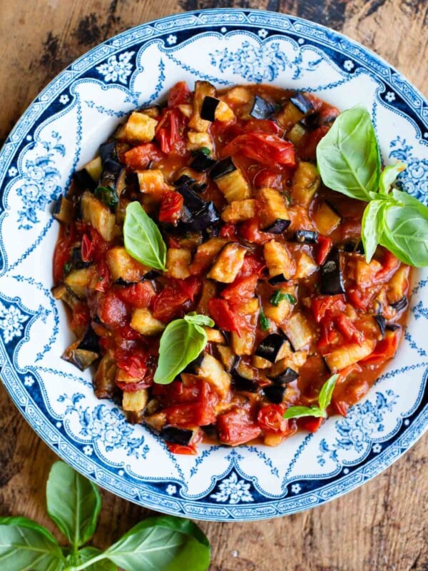 Fried eggplant in tomato sauce with fresh basil in a blue and white bowl on a wooden surface.
