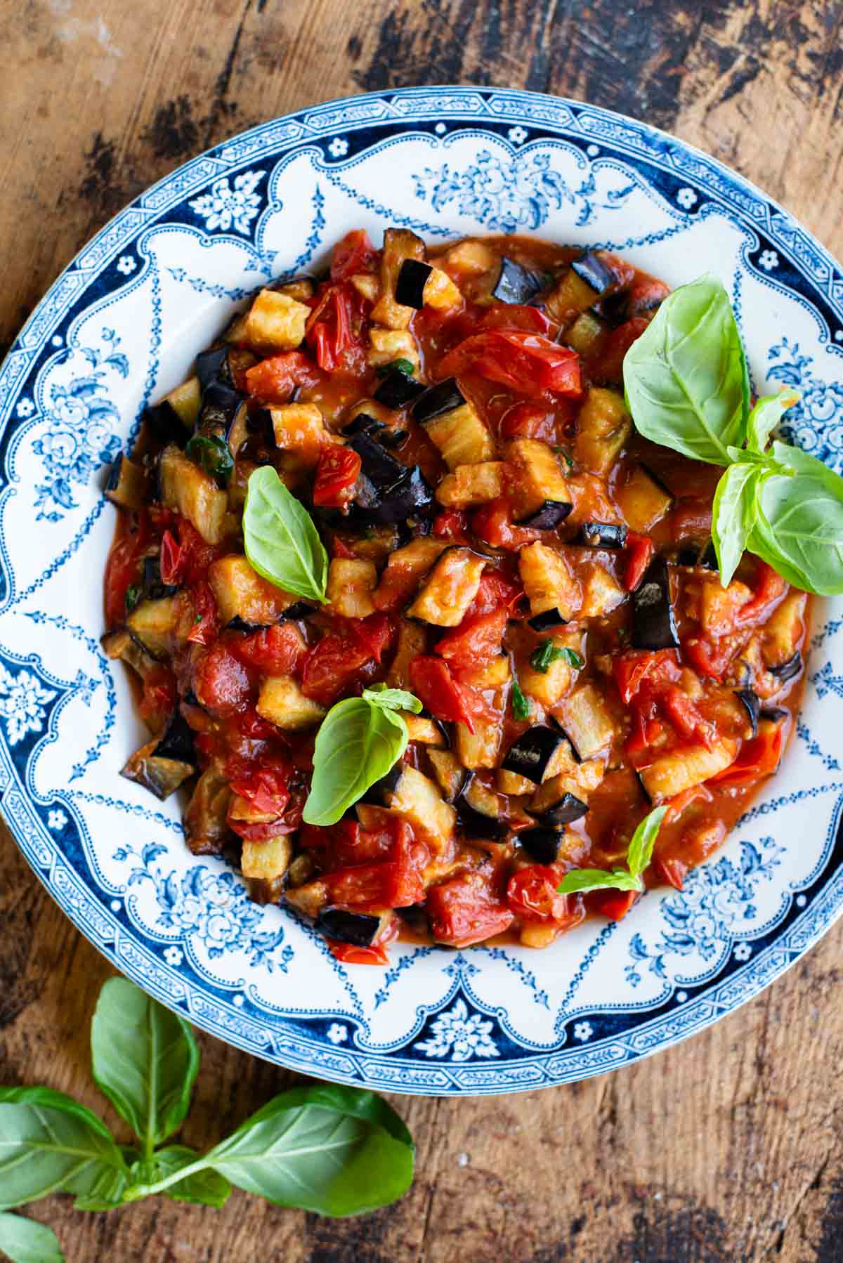 Fried eggplant in tomato sauce with fresh basil in a blue and white bowl on a wooden surface.