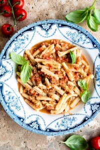 A blue and white vintage bowl with pasta in a fresh tomato sauce topped with basil. The bowl is on a travertine surface with cherry tomatoes and basil around.