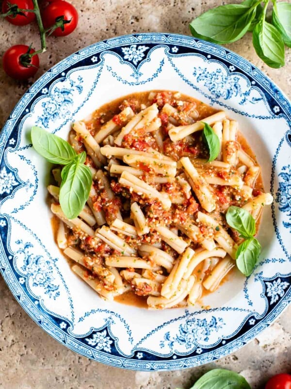 A blue and white vintage bowl with pasta in a fresh tomato sauce topped with basil. The bowl is on a travertine surface with cherry tomatoes and basil around.