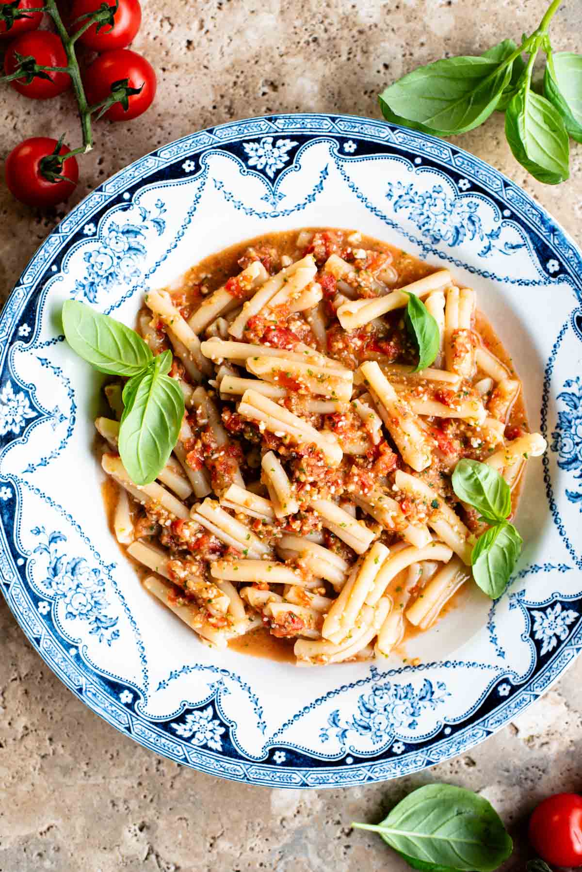 A blue and white vintage bowl with pasta in a fresh tomato sauce topped with basil. The bowl is on a travertine surface with cherry tomatoes and basil around.