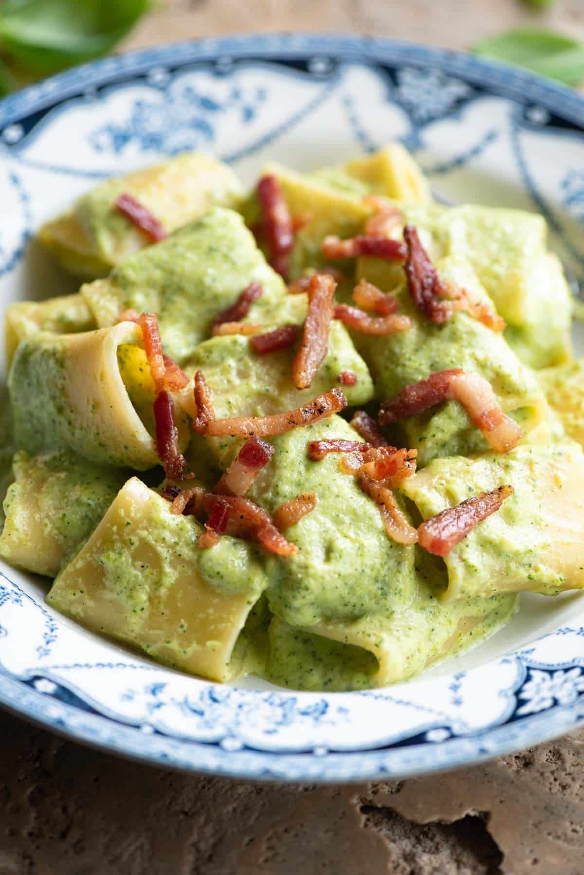 A side shot of paccheri pasta with a creamy zucchini sauce and topped with crispy pancetta. The bowl is blue and white.
