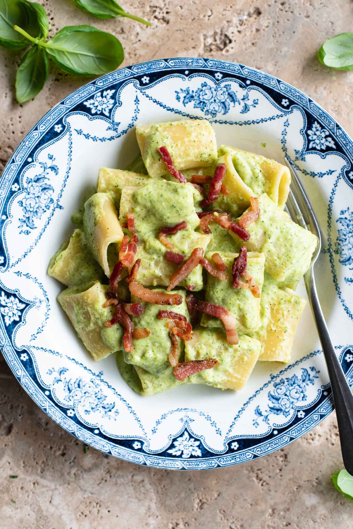 A creamy zucchini pasta in a blue and white bowl topped with crispy pancetta sitting on a travertine surface.