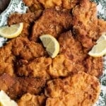 Pieces of crispy breaded pork tenderloin on a green and white serving platter with lemon wedges around.