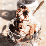 A glass dessert dish filled with a traditional Italian ice cream sundae. The sundae is topped with whipped cream, sour cherries, cocoa powder, and crumbled biscuits, with creamy gelato beneath. The Inside the Rustic Kitchen logo appears at the top.
