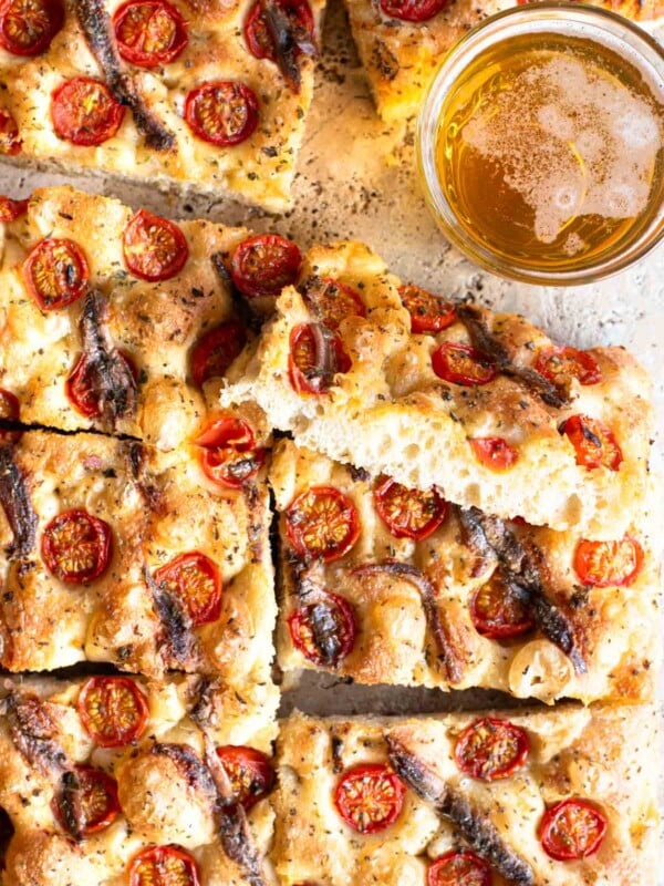 An overhead shot of a cherry tomato and anchovy focaccia cut into slices with a glass of beer at the side.