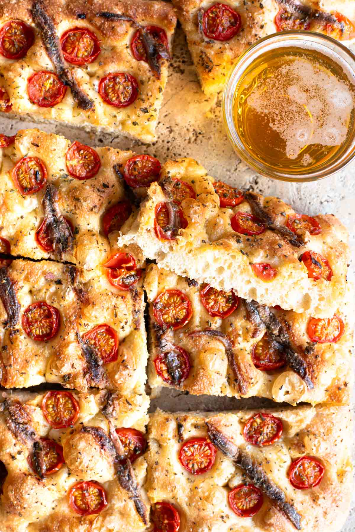 An overhead shot of a cherry tomato and anchovy focaccia cut into slices with a glass of beer at the side.