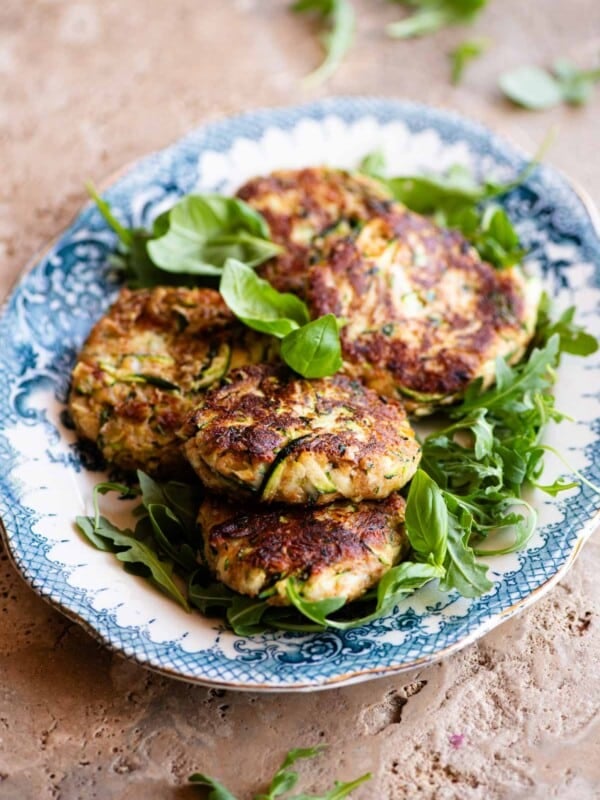 Five zucchini cakes (patties) sitting on top of arugula on a blue and white vintage style serving platter sitting on a travertine surface.