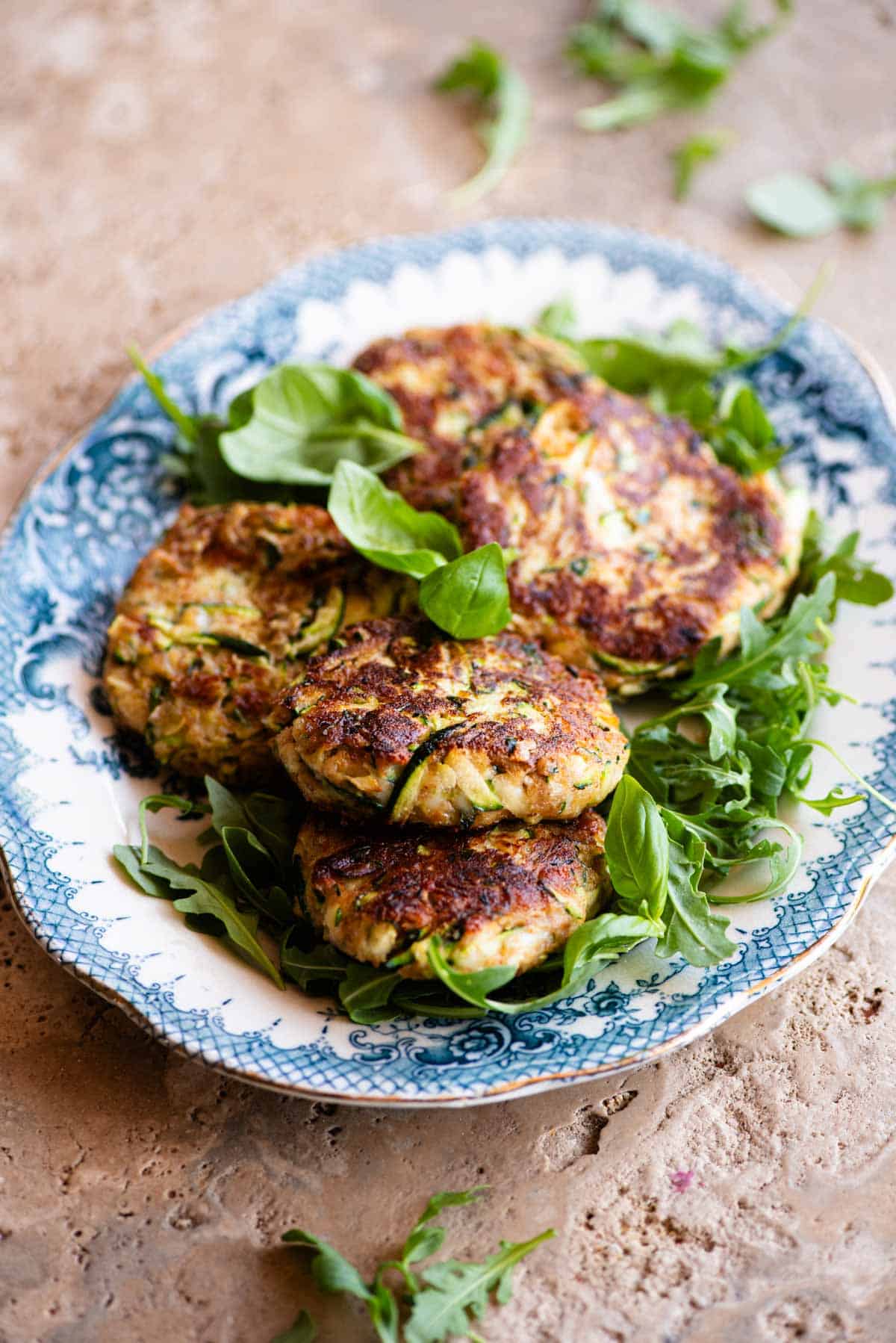 Five zucchini cakes (patties) sitting on top of arugula on a blue and white vintage style serving platter sitting on a travertine surface.