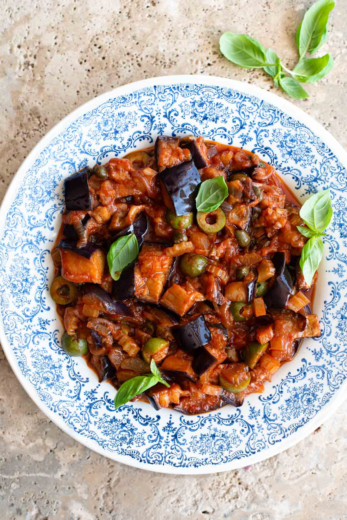 Sicilian caponata in a blue patterned bowl sitting on a travertine surface.
