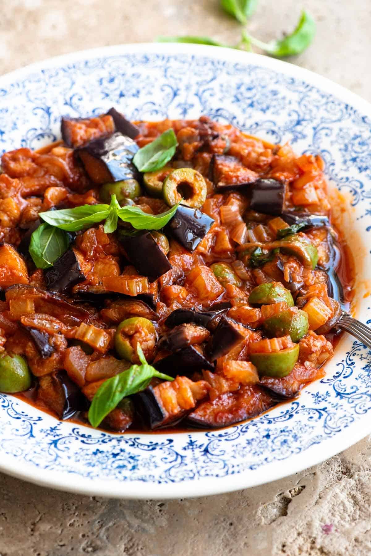 A side shot of caponata in a blue and white bowl topped with fresh basil leaves.