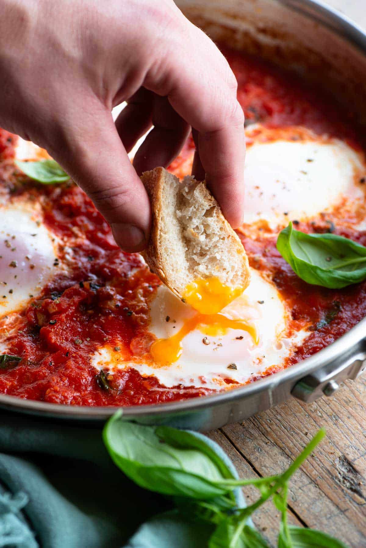 A hand dipping some bread into a poached egg with a runny yolk in a pan of tomato sauce.