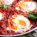 A close up of poached eggs in a tomato sauce with fresh basil in a stainless steel pan.