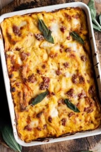 A whole butternut squash lasagna in a baking dish topped with sausage and sage leaves sitting on a wooden surface.