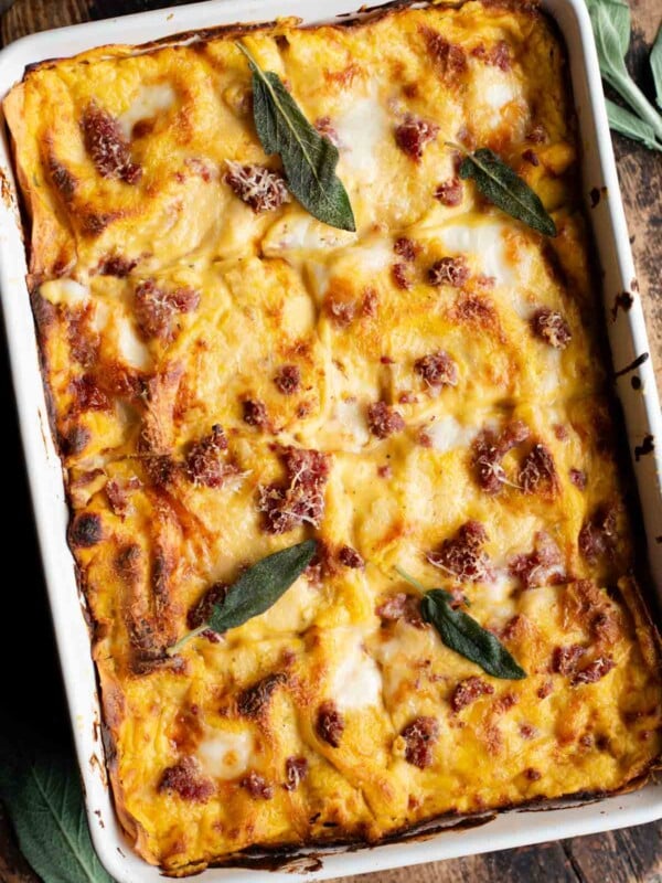 A whole butternut squash lasagna in a baking dish topped with sausage and sage leaves sitting on a wooden surface.