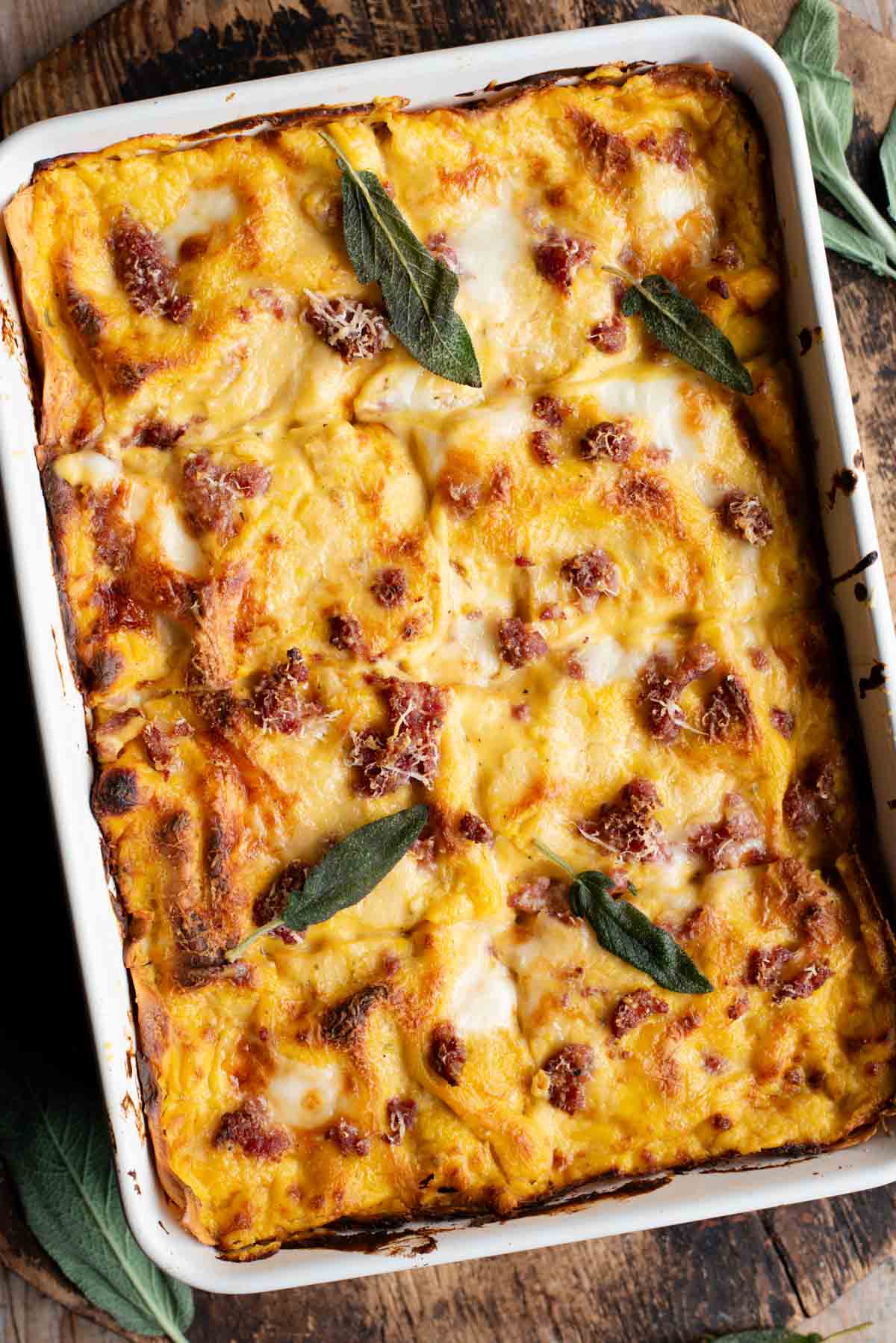 A whole butternut squash lasagna in a baking dish topped with sausage and sage leaves sitting on a wooden surface.