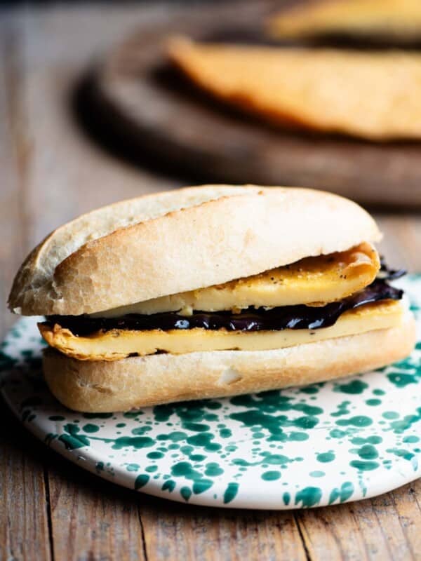 A cinque e cinque Livornese sandwich on a rustic wooden surface. The crusty roll is filled with chickpea farinata and marinated eggplant.