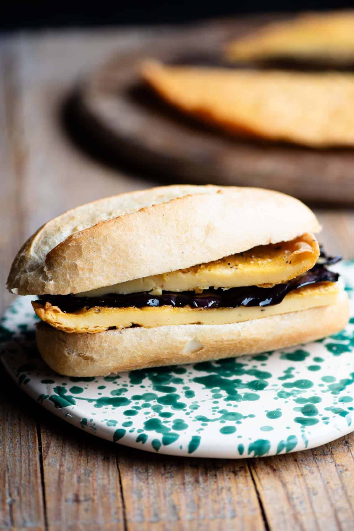 A cinque e cinque Livornese sandwich on a rustic wooden surface. The crusty roll is filled with chickpea farinata and marinated eggplant.