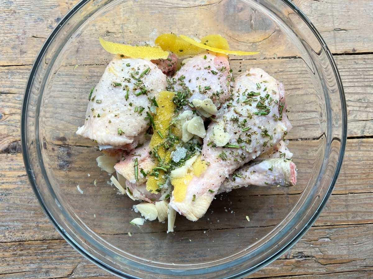 Chicken thighs and drumsticks in a glass bowl with lemon zest, herbs and garlic. It's sitting on a rustic wooden surface.