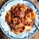 A bowl of pasta and hazelnut ragu on a wooden board with a glass of wine at the side.