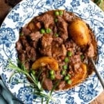 A close up of a beef stew with potatoes and peas in a blue and white bowl with rosemary.