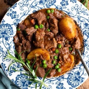 A close up of a beef stew with potatoes and peas in a blue and white bowl with rosemary.