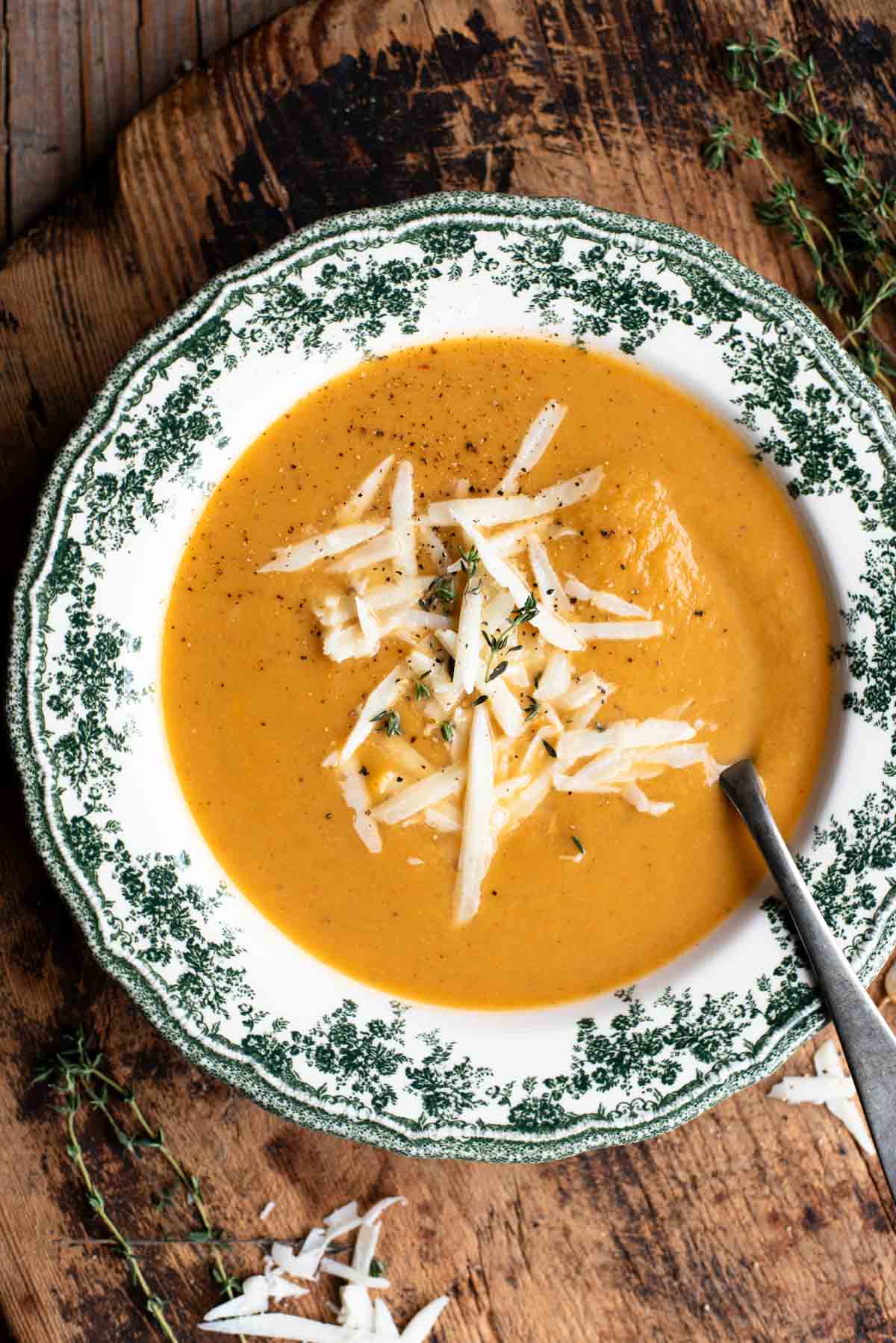 An overhead shot of a bowl of roasted vegetable soup in a green and white vintage bowl on a wooden board.
