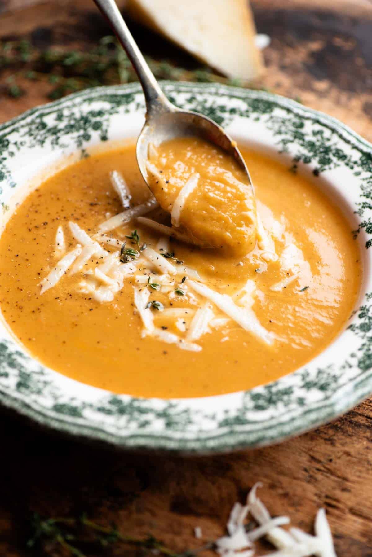 A close up of a spoon scooping up some roasted vegetable soup from a vintage style bowl.