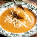 A close up of a spoon picking up some roasted vegetable soup from a green and white vintage style bowl.