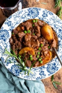 Italian beef stew with potatoes and peas in a blue and white floral bowl with rosemary and red wine at the side.
